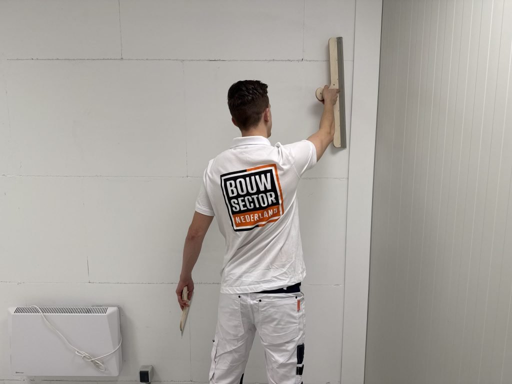 Stukadoor in witte werkkleding houdt de spaan op schouderhoogte tegen de wand om de pleister te verdelen